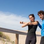 A man and woman in activewear stretch on a sunny Portugal beach boardwalk, promoting fitness and outdoor activities.