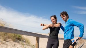 A man and woman in activewear stretch on a sunny Portugal beach boardwalk, promoting fitness and outdoor activities.
