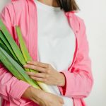 A woman in a pink jacket holding fresh spring onions against a white background.